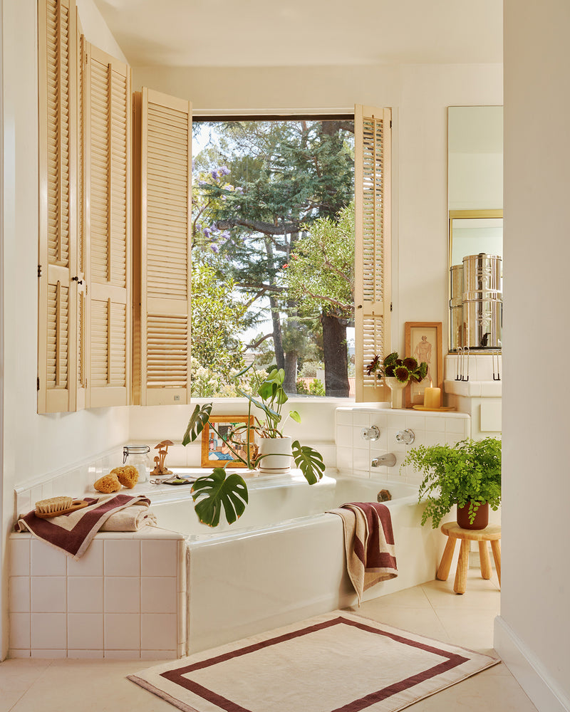Bathroom with a large window, bathtub, and plants with the two-toned bath towels in cacao and creme resting on bath tub.