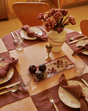 Set dining table with a terracotta linen tablecloth and cacao placemats and napkins, with floral arrangement, plates, glasses, and cutlery.  - view