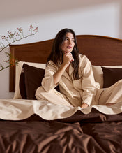 A woman in the Bed Threads Vanilla & Espresso Piped Long Sleeve Shirt stands barefoot by a bed with brown bedding in a sunlit room, near large windows overlooking greenery. - view