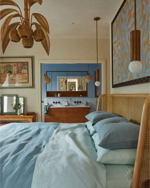 Cozy bedroom with a wooden bed frame, Bed Threads Coast 100% French Flax Linen Fitted Sheet in blue, pendant lights, rattan chandelier, large wall art, a framed picture, and a mirror above the wooden dresser reflecting the room.