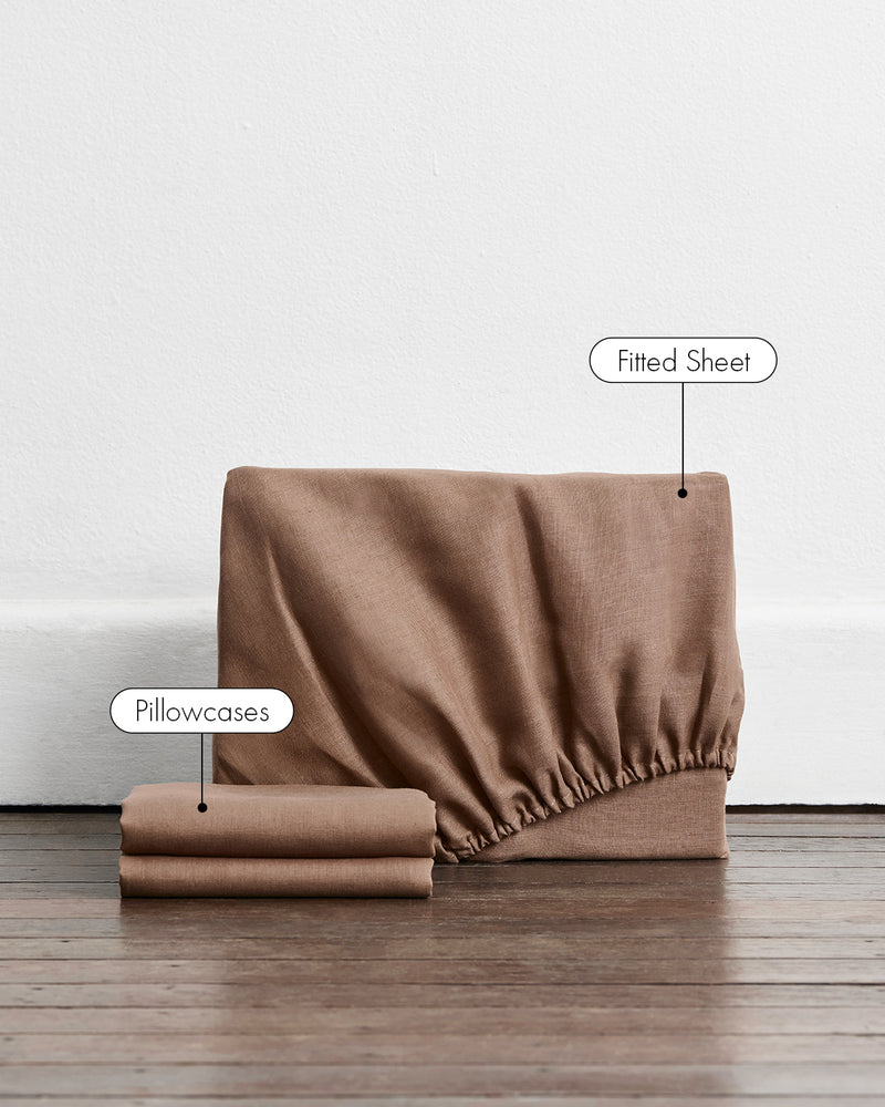 A 100% french flax linen fitted sheet and stacked pillowcase covers in a brown shade called portobello. White labels with black text point to the fitted sheet and pillowcases, set against a white wall and dark wood floor.