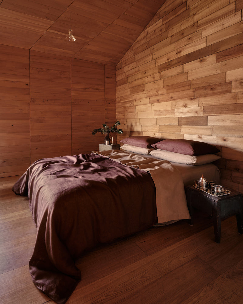 A cozy bedroom with a bed dressed in cacao and portobello 100% french flax linen bedding, set against a wall and ceiling made of warm wood paneling. A small bedside table holds a glass and a small decorative object.