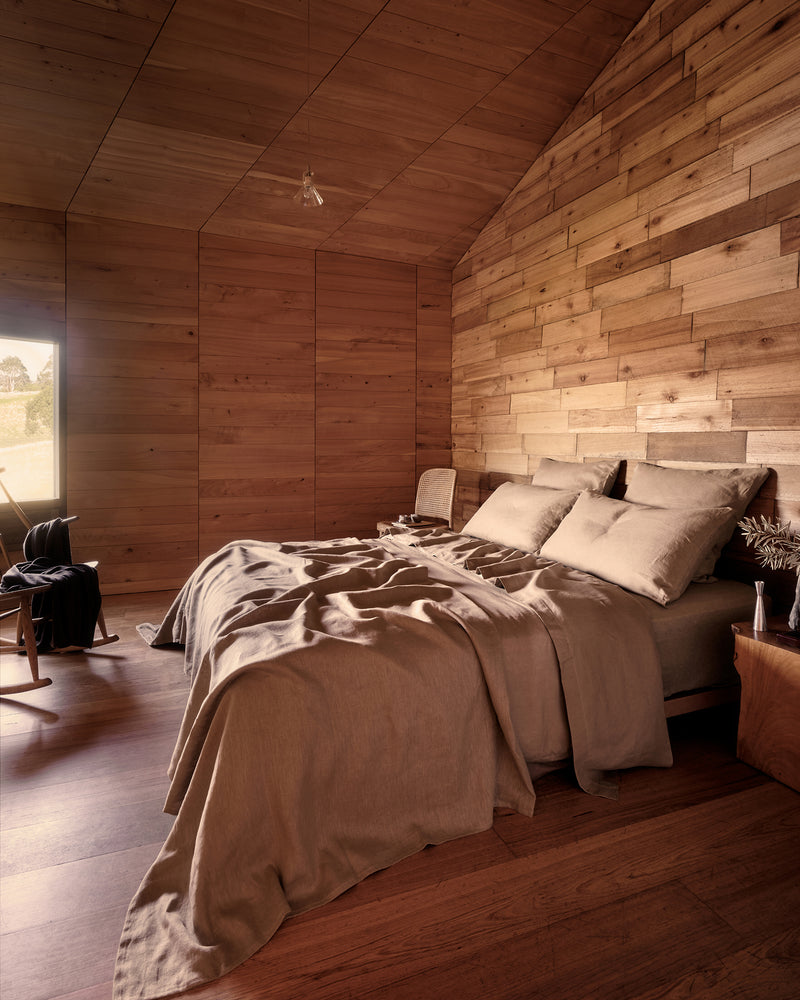 A rustic bedroom with a bed dressed in portobello 100% french flax linen, set against a wood-paneled wall and matching wood ceiling, with a window and chair visible on the left.