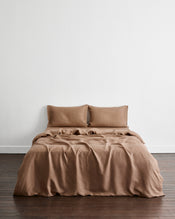 A bed dressed in a warm taupe-brown bedding set called Portobello in 100% french flax linen, including two pillowcases, a fitted sheet, and a duvet cover, shown in a minimalist room with a white wall and dark wood floor. - view