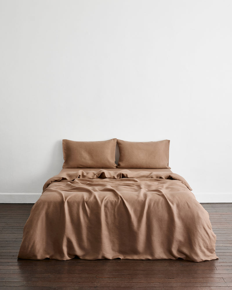 A bed dressed in a warm taupe-brown bedding set called Portobello in 100% french flax linen, including two pillowcases, a fitted sheet, and a duvet cover, shown in a minimalist room with a white wall and dark wood floor.