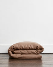 A folded brown-taupe duvet cover in 100% french flax linen, placed on a dark wood floor against a plain white wall. - view