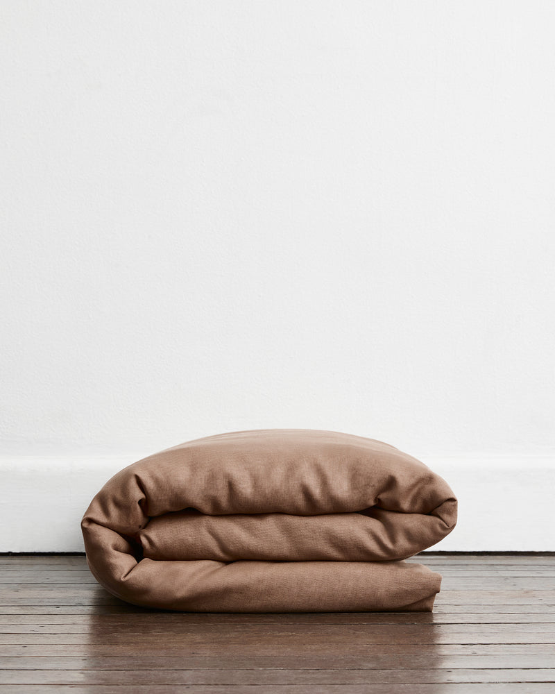 A folded brown-taupe duvet cover in 100% french flax linen, placed on a dark wood floor against a plain white wall.