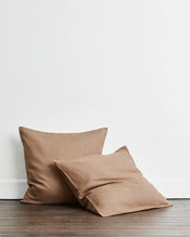 Two square european pillowcases in a warm taupe-brown 100% french flax linen fabric, one standing upright and one leaning in front, set on a dark wood floor against a plain white wall. - view