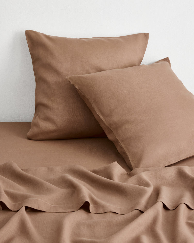 A close up image of two brown-taupe european pillowcases and sheets in 100% french flax linen, resting against a white wall. 