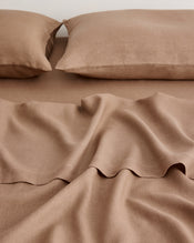 A close-up of a bed made with matching taupe-brown sheets and two pillowcases, showing soft linen fabric folds and a clean, coordinated look. - view