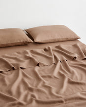 A bed dressed in a matching brown-taupe 100% french flax linen bedding set with two pillowcases and a sheet against a plain light background. - view