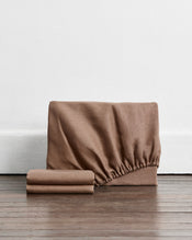 A 100% french flax linen fitted sheet and stacked pillowcase covers in a brown shade called portobello set against a white wall and dark wood floor.
 - view