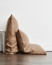 Two brown-taupe standard pillows in 100% french flax linen, one standing upright and one leaning against it, placed on a dark wood floor against a white wall. - view
