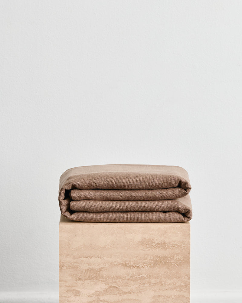 A folded warm brown throw in 100% french flax linen, folded neatly on a light beige stone pedestal against a plain white wall.