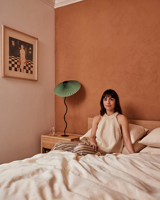 A woman with brown hair sits on a bed featuring the Bed Threads Crème 100% French Flax Linen Duvet Cover. A wooden nightstand with a green lamp and framed art stands against a peach wall, creating a cozy, inviting bedroom.