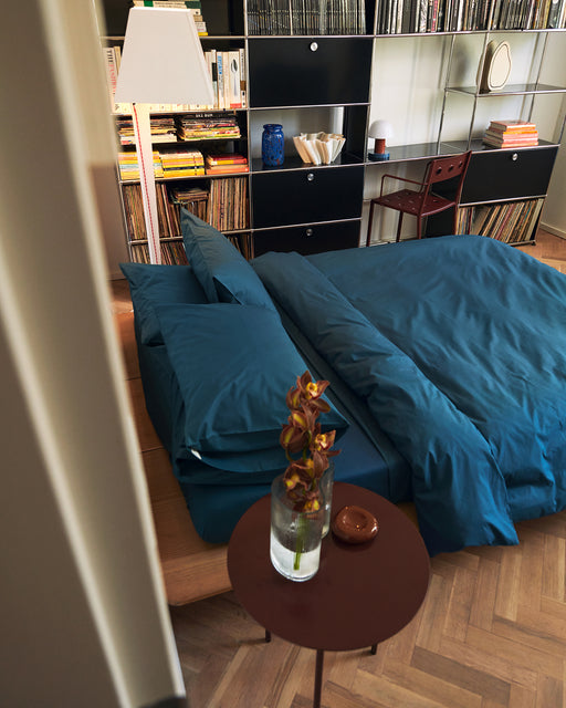 A cozy bedroom features a bed made up in hotel-quality bedding with the Bed Threads Aegean Organic Cotton Duvet Cover, beside a round table holding a vase of orange flowers. In the background are a bookshelf with books and records, a lamp, and a chair.