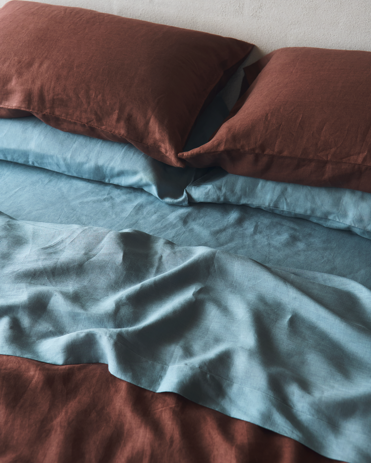 The Bed Threads Cacao & Lagoon 100% French Flax Linen Bedding Bundle features brown and blue tones with a smooth, softly wrinkled texture, creating a cozy and inviting look perfect for any bedroom.