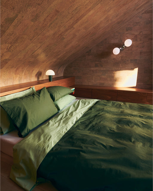 A cozy bedroom with a sloped wooden ceiling, a double bed made up in the Bed Threads Moss, Apple & Crisp White Organic Cotton Bedding Bundle, built-in wooden storage along the wall, and two modern lamps—one on a nightstand, one wall-mounted.