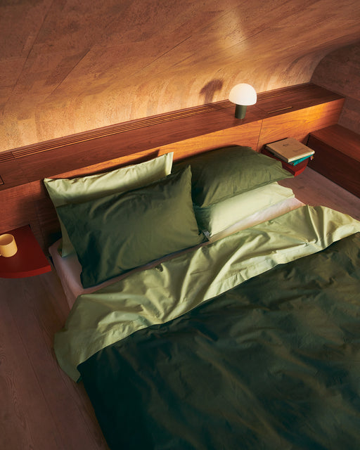 A cozy bed dressed in Bed Threads' Moss, Apple & Crisp White Organic Cotton Bedding Bundle sits by a wooden wall with a shelf holding a lamp, book, and mug. Warm lighting and organic cotton bedding create a relaxed, inviting atmosphere.
