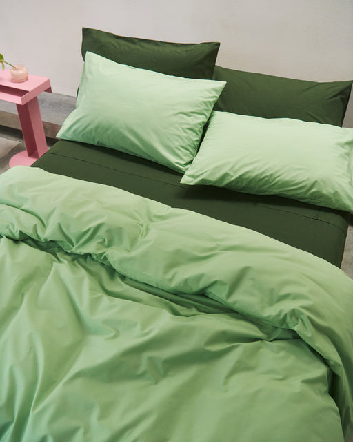 A neatly made bed features the Bed Threads Apple & Moss Organic Cotton Bedding Bundle, with dark green sheets, light green pillows, and a matching comforter made from GOTS certified organic cotton. A pink bedside table with a small plant sits in the corner.