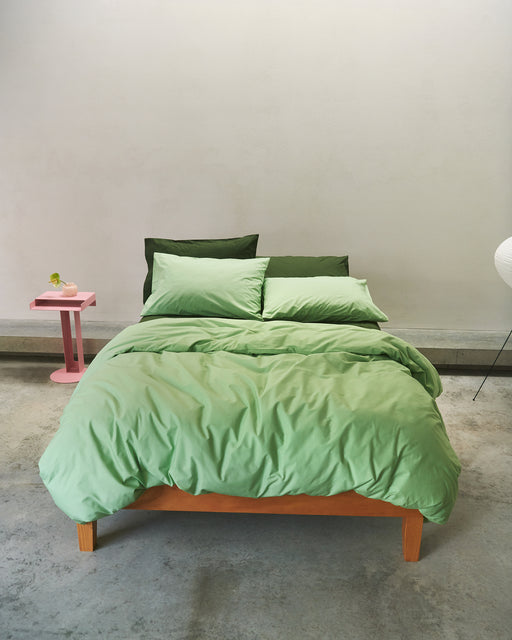 A neatly made bed with the Bed Threads Apple Organic Cotton Duvet Cover and two dark green pillows rests against a plain light wall. A pink side table with a small vase and minimal lamp stands beside the bed on a concrete floor.