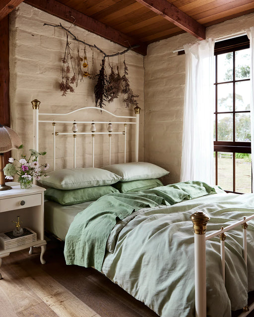 A cozy bedroom featuring a white metal bed frame, light green bedding topped with a Pistachio 100% French Flax Linen Flat Sheet by Bed Threads, a bedside table with flowers, dried plants above the bed, and sheer curtains on a large window.