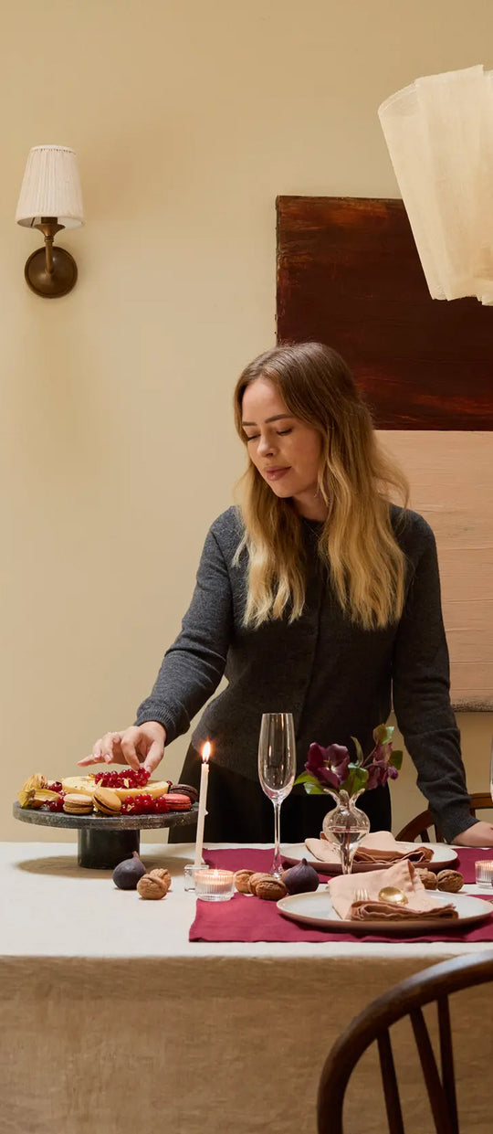 A woman with long blonde hair stands at a decorated dining table, reaching for a dessert on a cake stand. The table is set with candles, flowers, glassware, and plates, under warm indoor lighting.