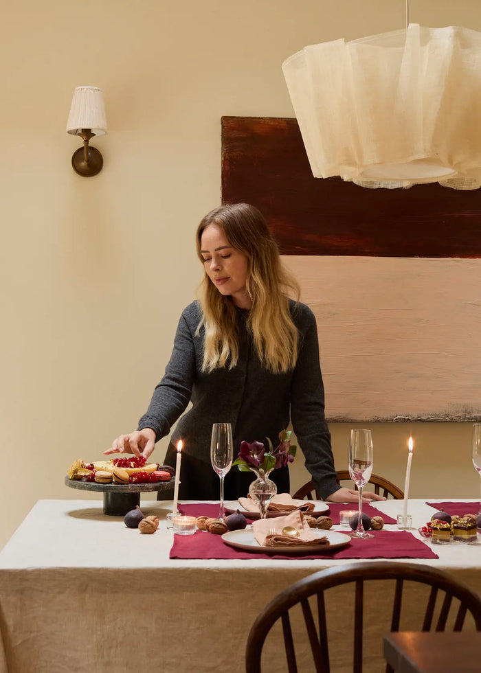 A woman stands at a dining table set with candles, flowers, pastries, and empty wine glasses. She arranges desserts on a cake stand under warm indoor light, with abstract art and neutral decor in the background.
