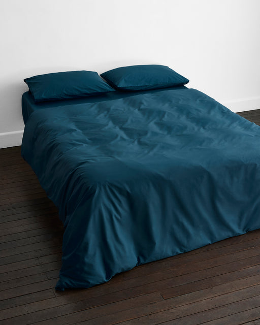 A neatly made bed features the Bed Threads Aegean Organic Cotton Duvet Cover in teal with two matching pillows, set on a dark wooden floor against a plain white wall.
