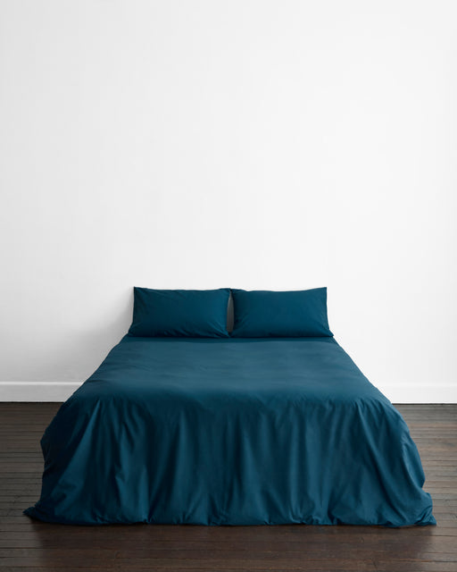 A minimalist room features a neatly made bed with the Aegean Organic Cotton Duvet Cover by Bed Threads, two matching pillows, a dark wooden floor, and a plain white wall.