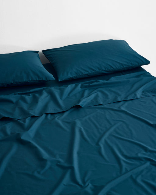 A neatly made bed with teal Bed Threads Aegean Organic Cotton King Pillowcases (Set of Two) against a plain white wall. The slightly wrinkled percale bedding creates a relaxed, inviting look.