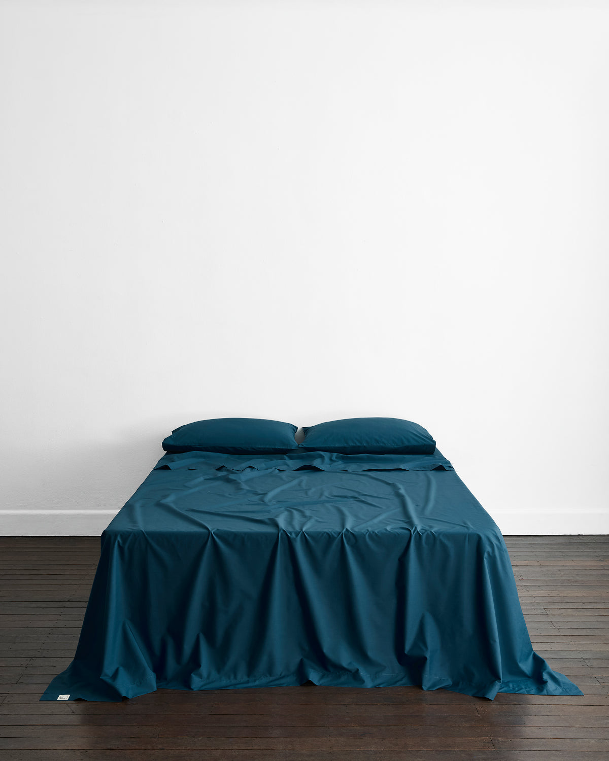 A neatly made bed with dark teal sheets and Bed Threads Aegean Organic Cotton King Pillowcases (Set of Two) stands against a white wall on a dark wooden floor in a minimalistic room.