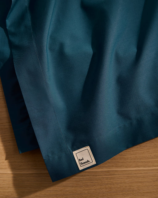 A close-up of the Bed Threads Aegean Organic Cotton Flat Sheet shows its dark teal percale fabric and small square label. The breathable, smooth bedding lies on a wooden surface, highlighting gentle folds and a subtle sheen.