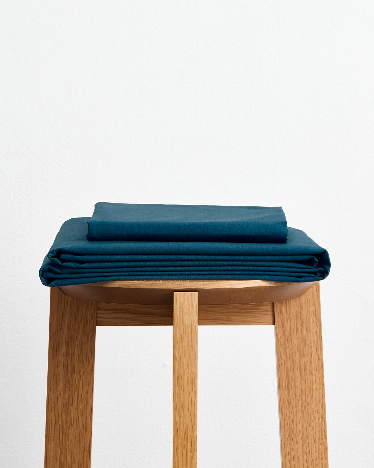 The Aegean Organic Cotton Sheet Set by Bed Threads, GOTS certified in teal, is neatly folded and stacked on a light wooden stool against a plain white background.