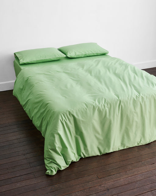A neatly made bed with the Bed Threads Apple Organic Cotton Duvet Cover and two matching pillows in light green, set against a white wall on a dark wooden floor.