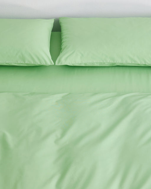 A neatly made bed features the Bed Threads Apple Organic Cotton Bedding Set with two matching pillows and a percale duvet cover, all in light green, displayed against a white wall.