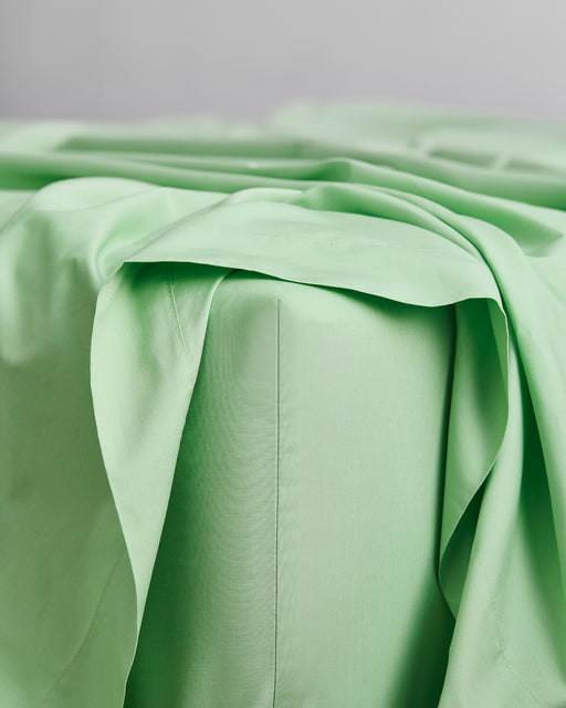 A close-up of a neatly made bed with the Apple Organic Cotton Fitted Sheet by Bed Threads in light green, and matching fabric draped over the corner, set against a plain, light-colored background.