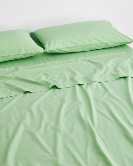 A neatly made bed with breathable Bed Threads Apple Organic Cotton Flat Sheet in light green, paired with two matching green pillows, set against a white wall.