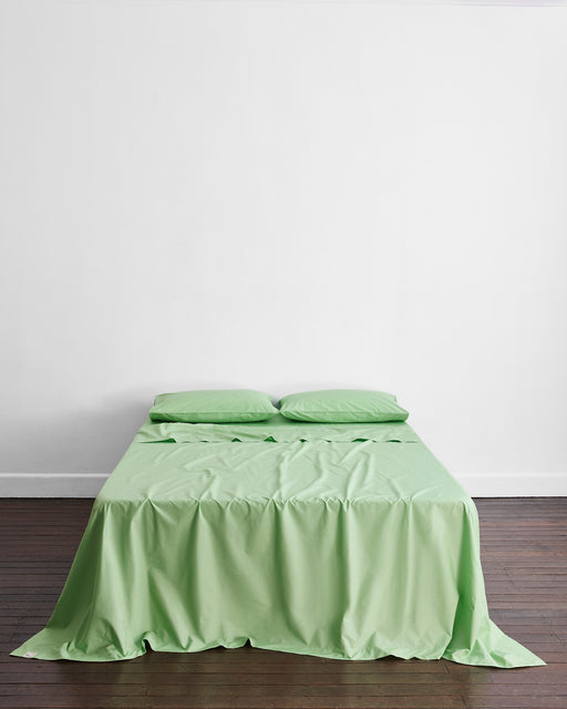 A neatly made bed features the Bed Threads Apple Organic Cotton Flat Sheet and two matching pillows, set against a white wall on a wooden floor, offering breathable GOTS certified comfort for a restful night's sleep.