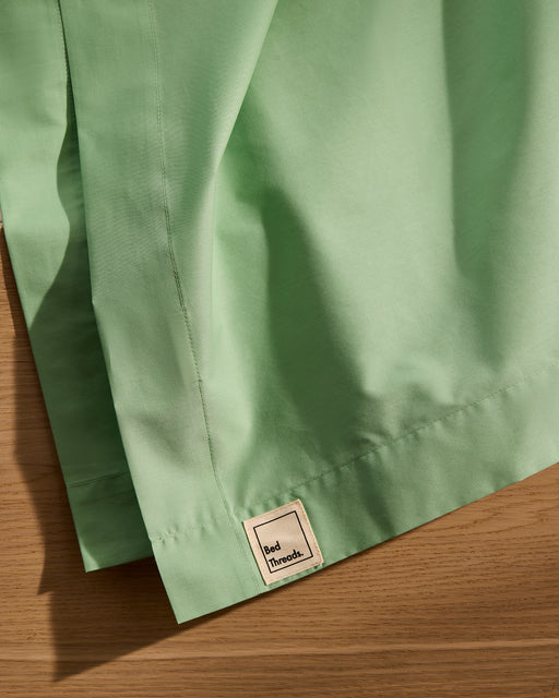 A close-up of the Apple Organic Cotton Flat Sheet by Bed Threads, in light green percale, draped on wood and displaying a small Bed Threads label.