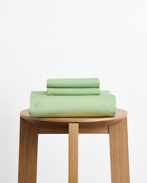 The Bed Threads Apple Organic Cotton Duvet Cover Set, in light green, is neatly stacked on a round wooden stool against a plain white wall.