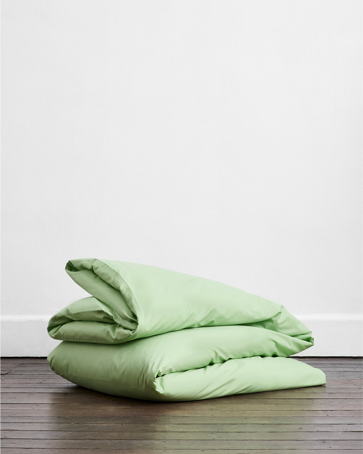 The Apple Organic Cotton Duvet Cover by Bed Threads, in light green, is neatly folded on a wooden floor against a plain white wall.