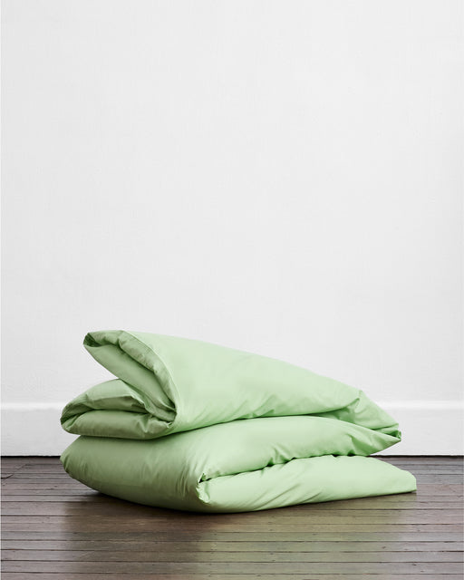 The Apple Organic Cotton Duvet Cover by Bed Threads, in light green, is neatly folded on a wooden floor against a plain white wall.
