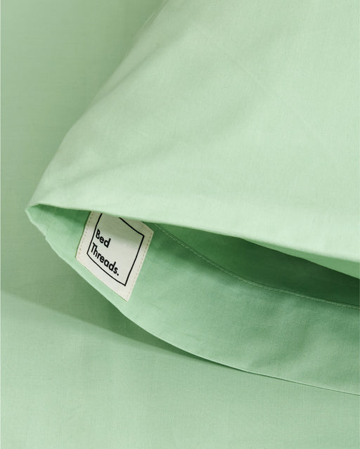 A close-up of Apple Organic Cotton Pillowcases (Set of Two) by Bed Threads, in light green percale, shows a smooth and breathable fabric with a visible white Bed Threads tag inside one pillowcase.