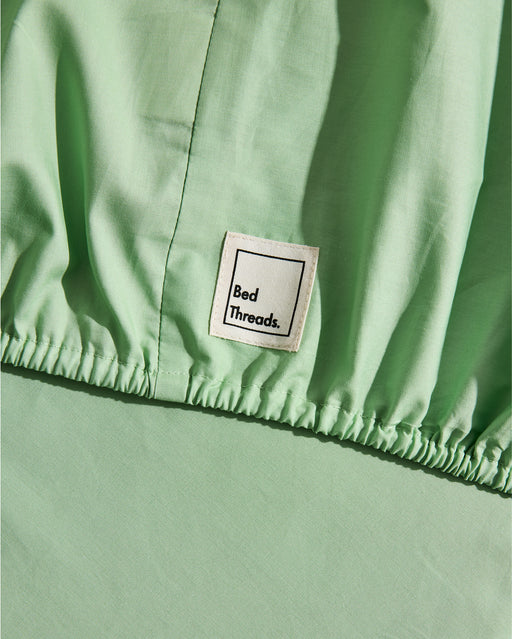 Close-up of the Apple Organic Cotton Fitted Sheet Set by Bed Threads, featuring a light green percale fabric, elastic edge, and a white label with "Bed Threads." in black text.