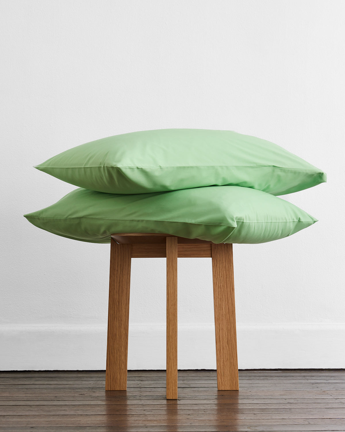 Two light green percale weave pillows from the Moss, Apple & Crisp White Organic Cotton Bedding Bundle by Bed Threads are stacked on a wooden stool, ideal for lovers of organic cotton bedding.