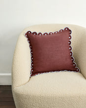 A cream armchair holds a maroon Bed Threads Cacao & Coast 100% French Flax Linen Arch Cushion Cover with scalloped edges and white trim, set against an off-white wall on a dark wooden floor. - view