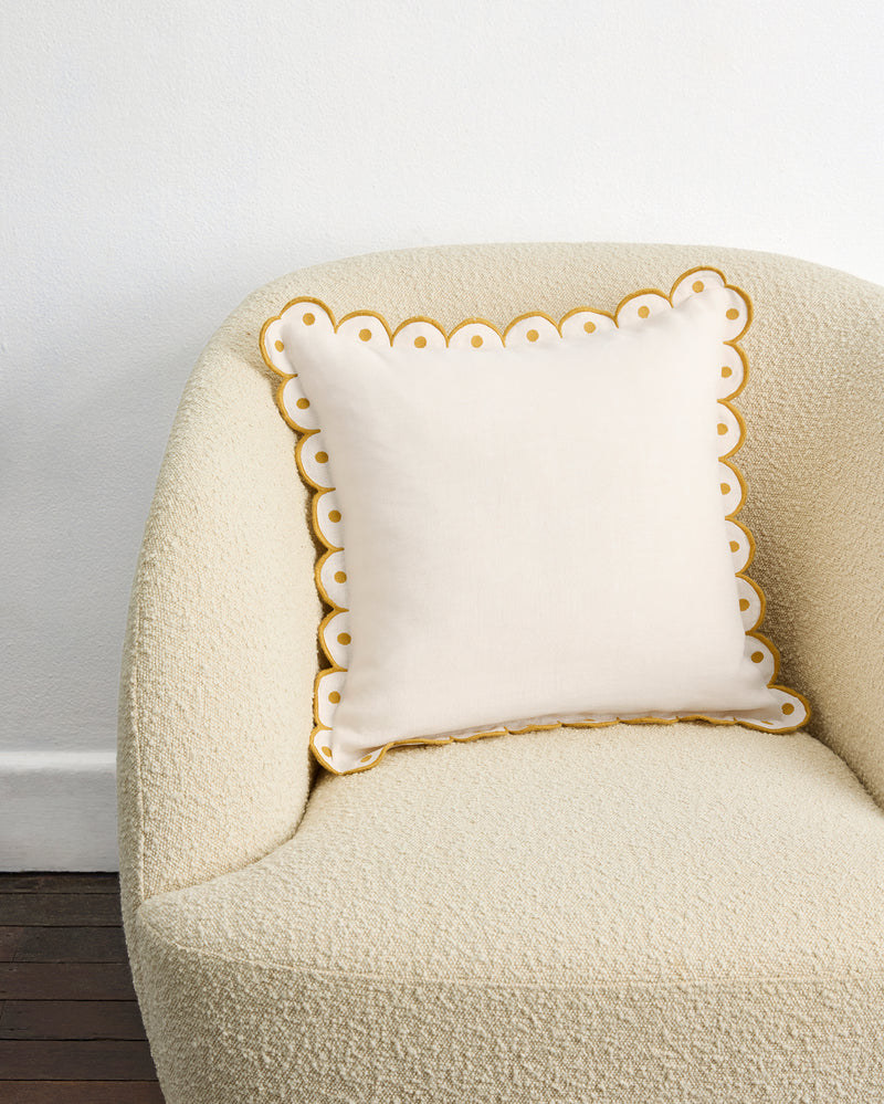 The Crème & Turmeric 100% French Flax Linen Arch Cushion Cover by Bed Threads, in cream with yellow trim and white dots, sits on a textured light beige armchair against a white wall and dark wooden floor.