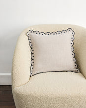 A cream armchair with a Bed Threads Oatmeal & Charcoal 100% French Flax Linen Arch Cushion Cover—featuring a scalloped edge and dark border—sits against a white wall on a wooden floor. - view