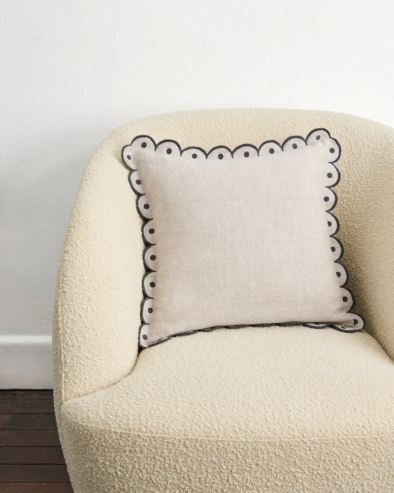 A cream armchair with a Bed Threads Oatmeal & Charcoal 100% French Flax Linen Arch Cushion Cover—featuring a scalloped edge and dark border—sits against a white wall on a wooden floor.
